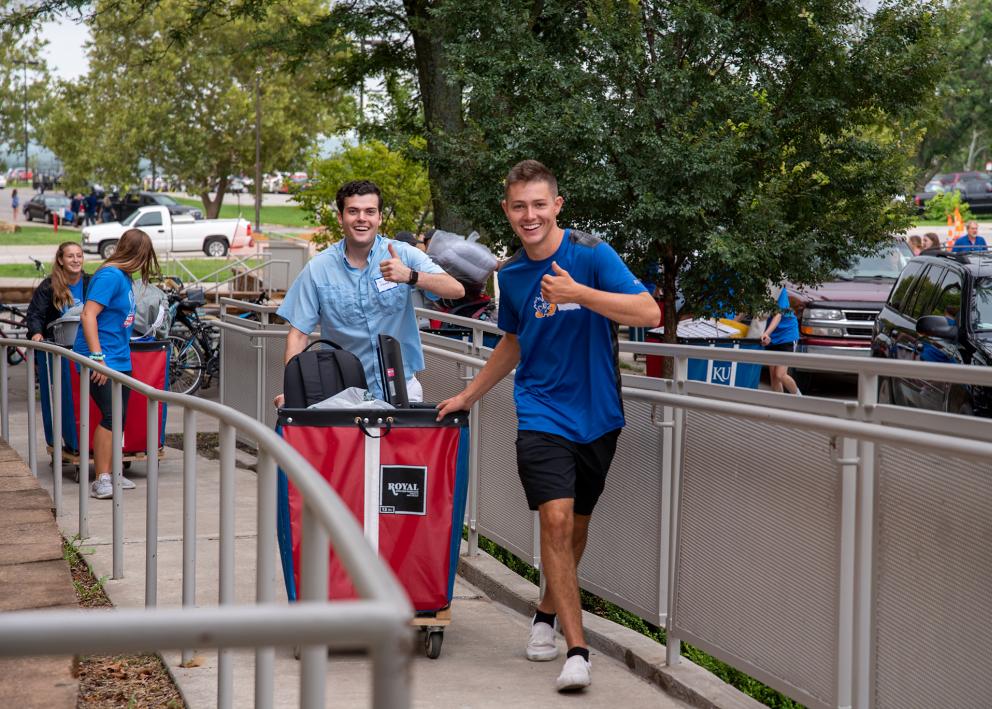 smiling new students pushing a cart up a ramp while giving an enthusiastic thumbs-up to the camera on move-in day