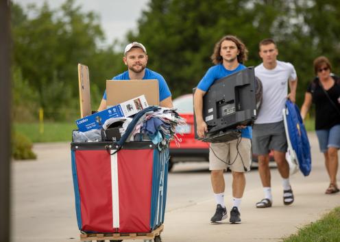 a line of student volunteers carrying and carting clothes, a TV, and a chair on move-in day