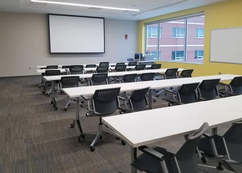 classroom interior with rows of tables and chairs, whiteboard, and front projector screen