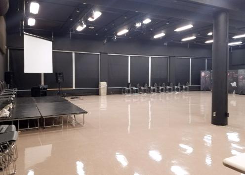 a large open theater room with a black out window wall, stage platforms, hanging projection screen, and many stackable chairs